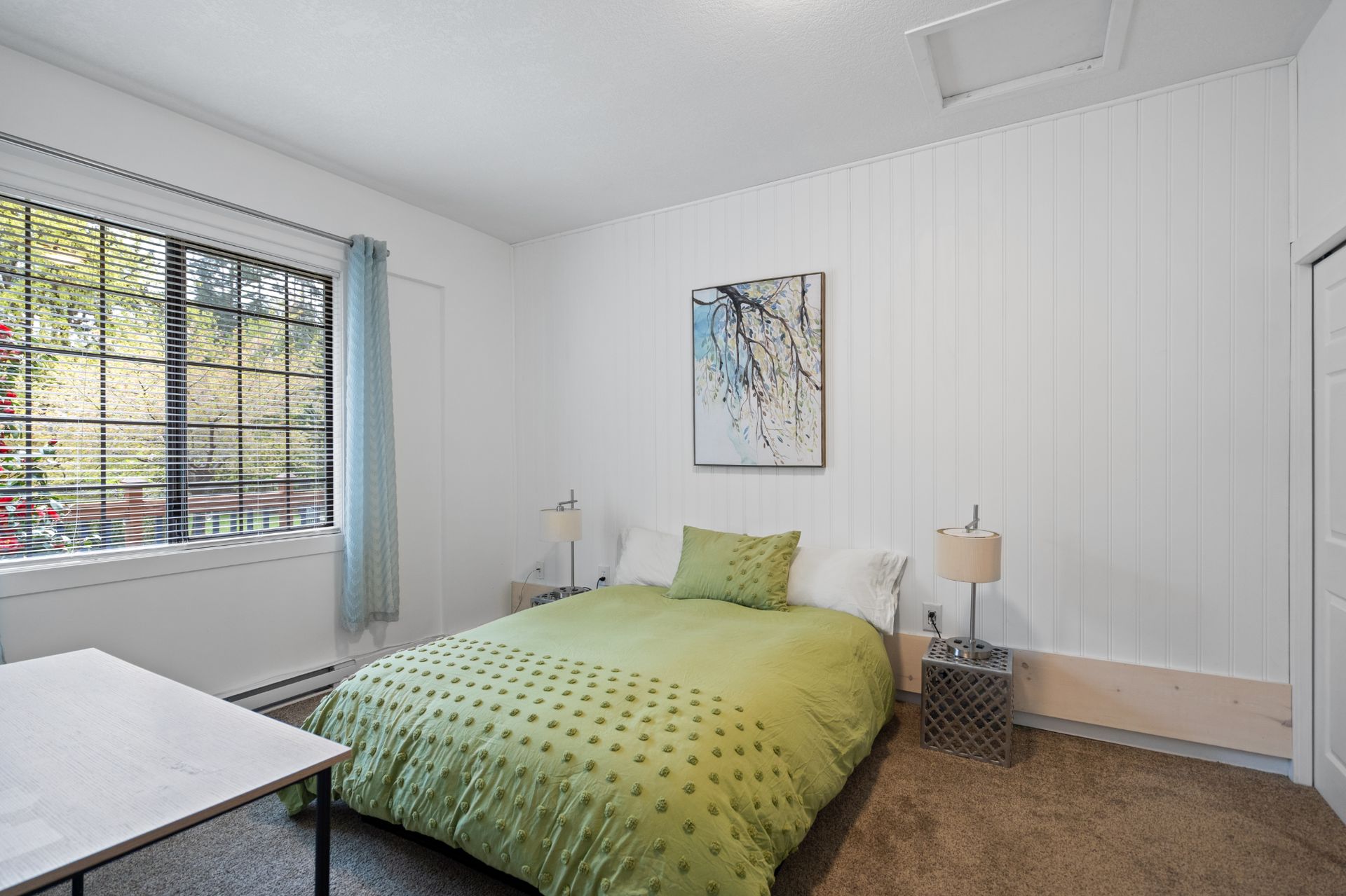Bright, minimalist bedroom with a queen bed featuring a green textured duvet, white paneled walls, matching bedside lamps on small tables, and a window with blinds and light blue curtains overlooking greenery.