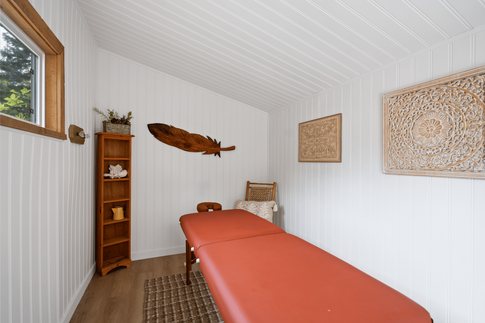 Cozy white treatment room with a massage table, wooden decor, and natural light from a window.